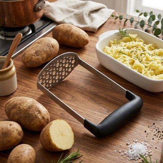 Presse purée manuel posé sur comptoir à côté de pommes de terre et d'une assiette de purée.